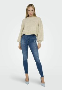 Pull en tricot couleur crème avec une coupe décontractée et un ourlet côtelé, associé à un jean bleu fitted avec des détails déchirés et une longueur cheville.