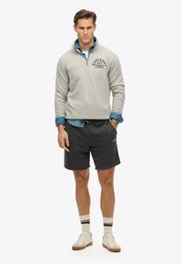 Lichtgrijze fleece hoodie met een ritskraag en een logo op de borst, gedragen over een blauwe denimshirt en gecombineerd met zwarte shorts en crèmekleurige sneakers.