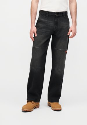 Mann trägt lockere schwarze Jeans mit Knie-Patches und braune Arbeitsschuhe, steht vor einem schlichten hellen Hintergrund.