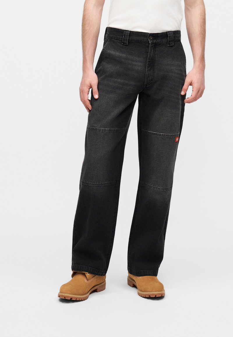 Άνδρας που φοράει φαρδιά μαύρα τζιν με επιθέματα στα γόνατα και καφέ εργοστασιακά μποτάκια, στέκεται μπροστά σε απλό φωτεινό φόντο.