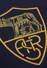 Marine stof met een getextureerd, geel geborduurd schildlogo met een beer en figuren, waardoor ingewikkelde details en een gedurfde kleurcontrast opvallen.