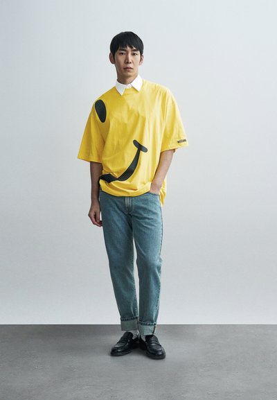 Κίτρινο φαρδύ T-shirt με μαύρο γραφικό χαμογελαστό πρόσωπο, συνδυασμένο με ανοιχτό μπλε τζιν και μαύρα μοκασίνια. Ορατή λευκή λεπτομέρεια στον γιακά.