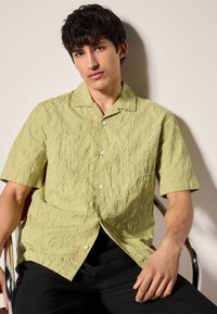 Korte mouwen lichtgroen shirt met getextureerde cirkelpatronen, knoopsluiting en een brede kraag, gecombineerd met een zwarte broek.