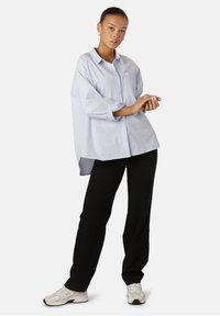 Camicia oversize azzurro chiaro con bottone e tasca, maniche lunghe arrotolate ai polsini, abbinata a pantaloni neri e sneakers bianche.