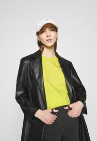 Blazer in pelle nera sopra una maglietta gialla brillante con logo, abbinato a pantaloni neri. Cappellino da baseball bianco con logo ricamato, vista ravvicinata.
