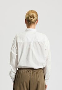 Camicia bianca con colletto classico, maniche lunghe e vestibilità comoda, abbinata a pantaloni marroni con cintura elastica.