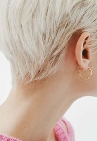 Aro de oro retorcido colocado en un peinado corto de cabello rubio claro. El fondo es blanco liso, con un suéter de un suave color rosa visible.