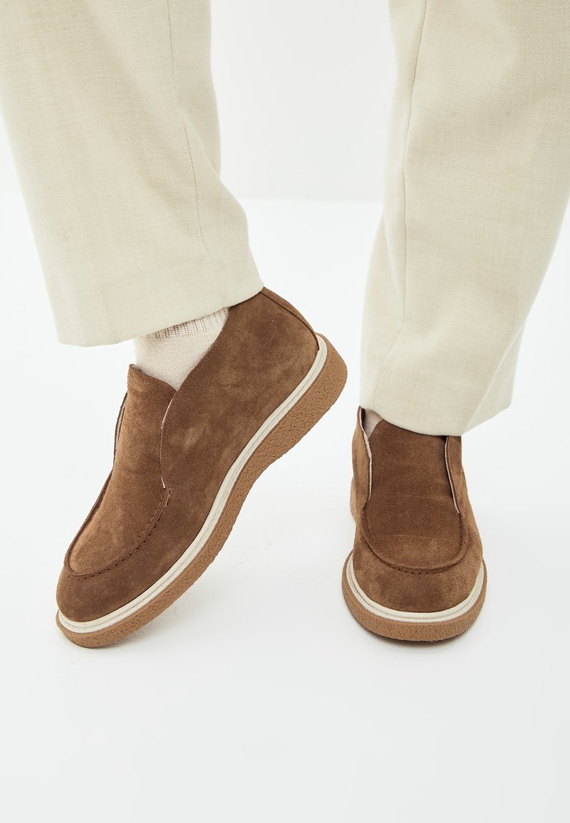 Chaussures en daim marron sans lacets portées avec un pantalon beige clair et des chaussettes crème, sur fond blanc.