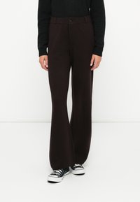 Personne portant un pantalon large marron foncé avec fermeture à bouton et à zip, un haut noir à manches longues, et des baskets basses noir et blanc.