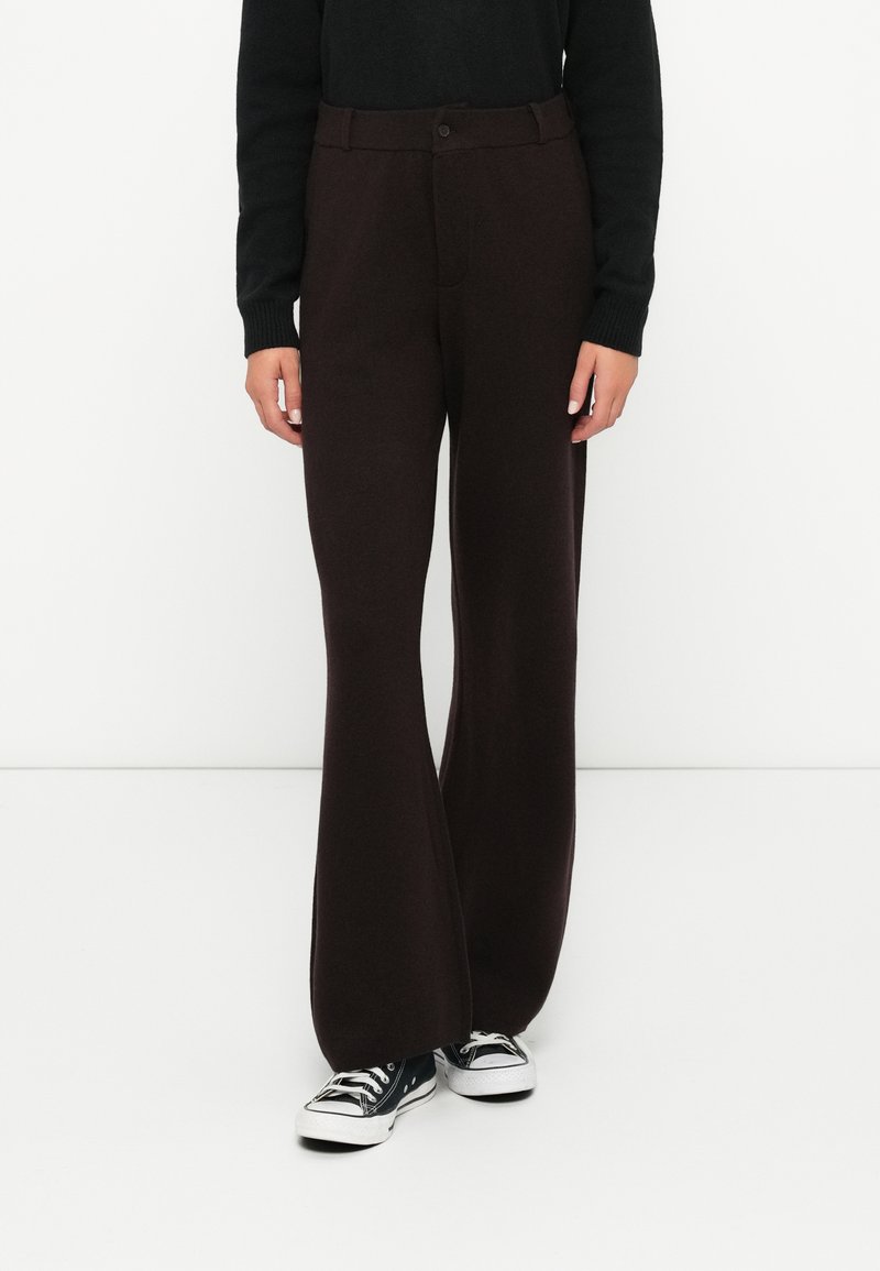 Personne portant un pantalon large marron foncé avec fermeture à bouton et à zip, un haut noir à manches longues, et des baskets basses noir et blanc.