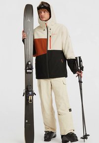 Ski-outfit met een kleurgeblokte jas in crème, zwart en oranje, beige broek, zwarte ski's en zwarte skistokken met textuurrijke oppervlakken.