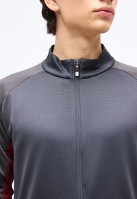 Άτομο που φοράει φερμουάρ γκρι και σκούρο γκρι μπουφάν με μπορντό λεπτομέρειες, με γυμνό λαιμό και κάτω μέρος του προσώπου απέναντι σε λευκό φόντο.
