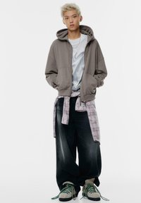 Un giovane con capelli biondi corti indossa un oversized grigio, una camicia bianca, una camicia a quadri legata in vita, pantaloni neri larghi e scarponi da trekking.