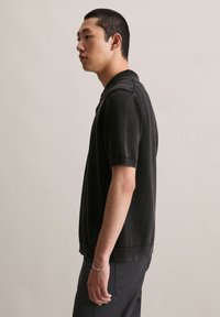 Młody mężczyzna w czarnej polówce z krótkim rękawem i ciemnych spodniach, stojący w profilu na jednolitym jasnym tle.