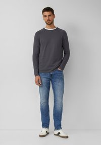 Mężczyzna stojący przy szarej ścianie, ubrany w ciemnoszary longsleeve, niebieskie dżinsy i białe tenisówki z czarnymi paskami.