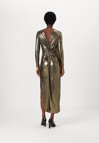 Zlaté metalické šaty s dlouhým rukávem, hlubokým výstřihem na zádech, svinutým detailem v pase a bočními rozparky. Nosené s černými vysokými podpatky.