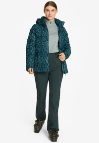 Chaqueta de plumas de color teal con un patrón texturizado, capucha ajustada y bolsillos con cremallera, llevada sobre una camiseta gris claro y pantalones verde oscuro con cubrebotas.
