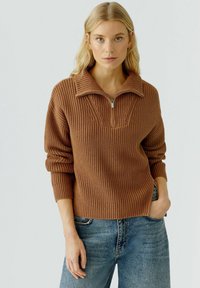 Bruine gebreide trui met een halve rits kraag, ribbeltextuur en kabelgebreide accenten. Draagt met lichtblauwe denim jeans.