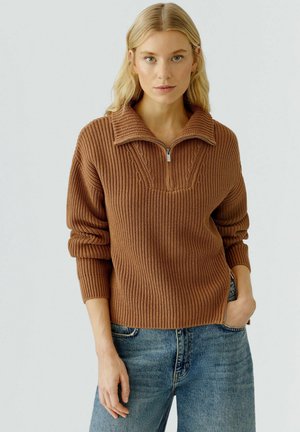 Bruine gebreide trui met een halve rits kraag, ribbeltextuur en kabelgebreide accenten. Draagt met lichtblauwe denim jeans.