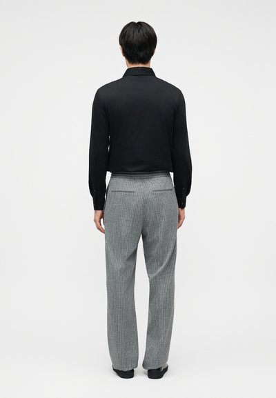 Άνθρωπος με το ανάστημά του να φαίνεται, φοριέται μαύρο πουκάμισο μακρυμάνικο και γκρι παντελόνι με ριγέ λεπτομέρειες, στέκεται μπροστά σε ένα απλό ανοιχτόχρωμο φόντο.