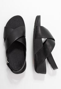 Ett par svarta sandaler med breda korsade läderremmar och strukturerade fotbäddar, visade från ovan- och sidovy på en vit bakgrund.