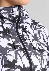 Chaqueta con cremallera que presenta un patrón de hojas de palma en blanco y negro, con un alto cuello, capucha ajustable y logo en el pecho.