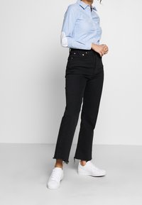 Chemise bleu clair à boutons avec des coudières blanches à pois, associée à un jean noir taille haute et des baskets blanches.