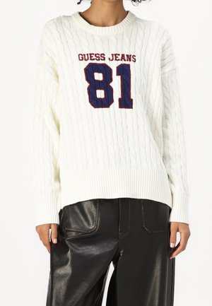 Bílý svetr s kabelovým vzorem s nápisem "GUESS JEANS 81" v červené a námořnické modři, nošený s černými koženými kalhotami na stojící osobě.