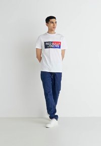 Balta T-kreklā ar daudzkrāsainu "RED BULL RACING" logotipu; apvienota ar zilu džinsu biksēm un baltām sporta apavu kedām, stāvot uz neitrāla fona.