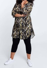 Button-Up-Tunika im mehrfarbigen abstrakten Muster mit Grün, Schwarz und Weiß. Mit 3/4-Ärmeln und abgerundetem Saum, kombiniert mit schwarzen Caprihosen.