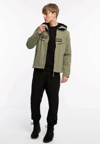 Olijfgroene jas met voorzakken, ritsluiting en donkerblauwe capuchon. Gecombineerd met zwarte broek en zwarte veterschoenen.