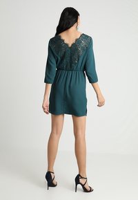 TAPESTRÍNDES Kleid mit 3/4-Ärmeln und einem Spitzenrücken-Design, mit tailliertem Schnitt und einer Länge über dem Knie, ergänzt durch schwarze Absätze.