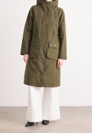 Persoon die een olijfgroene knielange jas met grote voorkanten zakken draagt, gecombineerd met wijde witte broek en zwarte spitse schoenen.