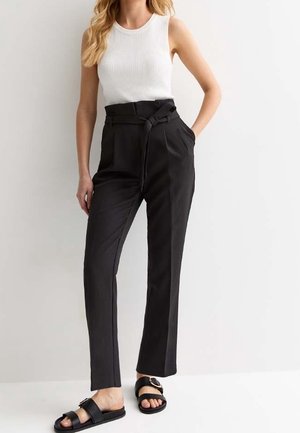 Femme portant un haut blanc sans manches côtelé, un pantalon noir taille haute avec ceinture, et des sandales slides noires à boucles.