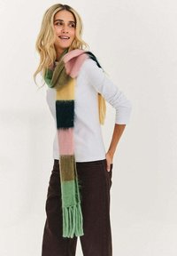 Kobieta w długim, wielokolorowym szaliku w paski, białej bluzce z długim rękawem i ciemnych spodniach, uśmiechająca się i patrząca w bok.