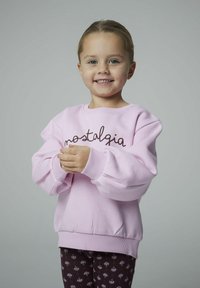 Giovane ragazza che sorride, indossando una felpa rosa chiaro con la scritta "nostalgia" e pantaloni scuri con motivo, in piedi su uno sfondo grigio uniforme.