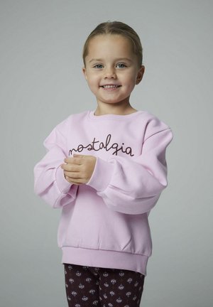 Niña sonriente, vestida con un sudadera rosa claro con el texto "nostalgia" y pantalones oscuros estampados, de pie frente a un fondo gris liso.