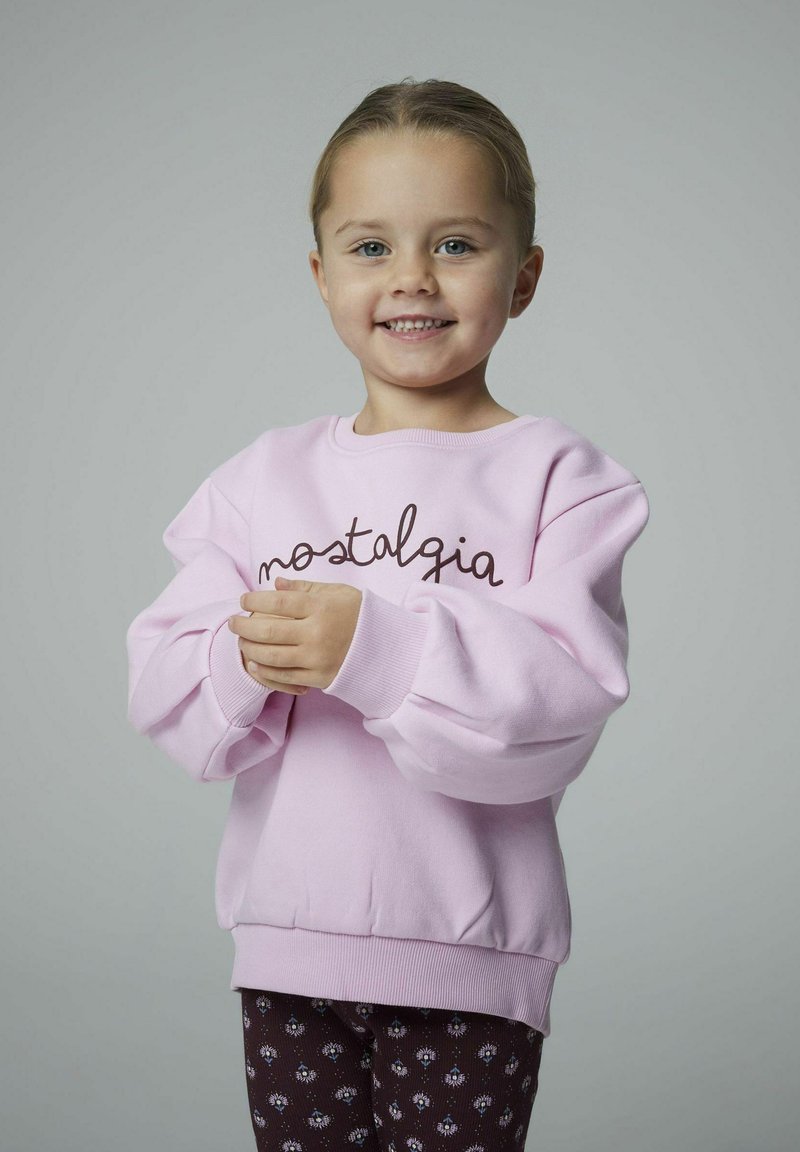 Giovane ragazza che sorride, indossando una felpa rosa chiaro con la scritta "nostalgia" e pantaloni scuri con motivo, in piedi su uno sfondo grigio uniforme.