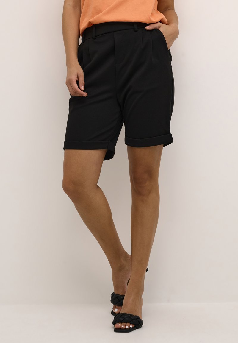 Shorts noirs ajustés avec des revers, fabriqués en tissu léger. Assortis avec un haut orange et des sandales à talons tressées noires.