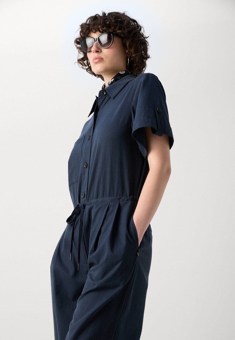 Frau mit lockigem Haar, die schwarze Sonnenbrille trägt und einen kurzärmeligen, marineblauen Overall anhat, mit den Händen in den Taschen, stehend vor einem weißen Hintergrund.