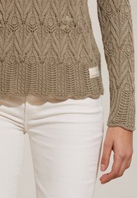Maglione lavorato a maglia beige con orlo scallop, motivo intricato e piccolo dettaglio floreale. Abbinato a jeans bianchi aderenti.