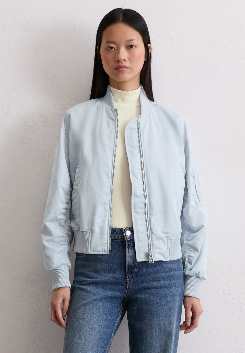 Jasnoniebieska kurtka bomberka o gładkiej fakturze, z ściągaczami przy mankietach i zamkiem błyskawicznym. Nosi się ją na kremowy sweter typu golf i niebieskie dżinsy.