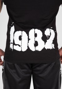 Svart bomullströja med stor vit sliten tryck av siffran "1982" på ryggen, i kombination med svarta mesh-shorts.