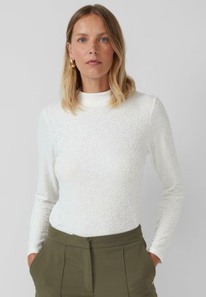 Blonde vrouw met blauwe ogen, die gouden oorbellen, een witte textuur lange mouw top en olijfgroene broek draagt, met haar handen in haar zakken.