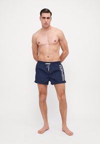 Tumedad ujumisshortsid valge paelaga ja külglogoga. Kiire kuivamisega kangas, keskpõlve pikkus, elastne vöökoht, kerge disain.