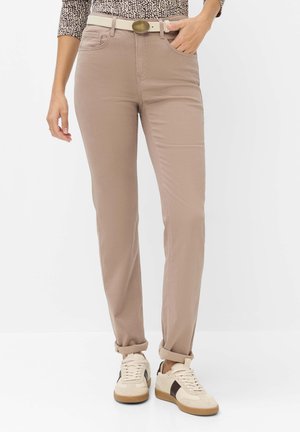 Beige broek die bij de enkels is opgerold, gedragen met een witte riem, een top met patroon en witte sneakers met zwarte accenten. Eén hand in de zak, één ernaast.