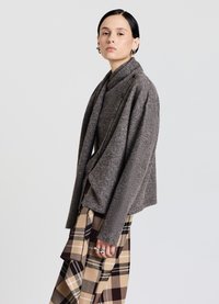 Cardigan in lana grigia con collo a scialle, caratterizzato da bottoni, indossato sopra una gonna a quadri beige e marrone. Modella in posa di profilo.