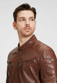 Chaqueta de cuero marrón con cuello alto, dos bolsillos de pecho con cremallera, detalles acolchados en los hombros y una textura suave.