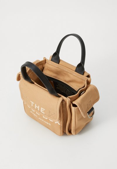 Beige canvas handtas met zwarte leren hengsels, voorzien van voorzakken en geperst text. Compact rechthoekig formaat met een gestructureerde afwerking.