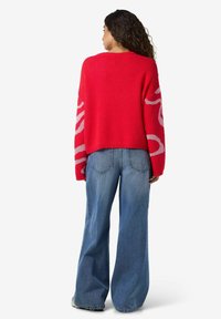 Rdeč pleten pulover z abstraktnim vzorcem svetlo sive barve na rokavih, kombiniran z ohlapnimi modrimi kavbojkami. Mehka tekstura in krojen dizajn.